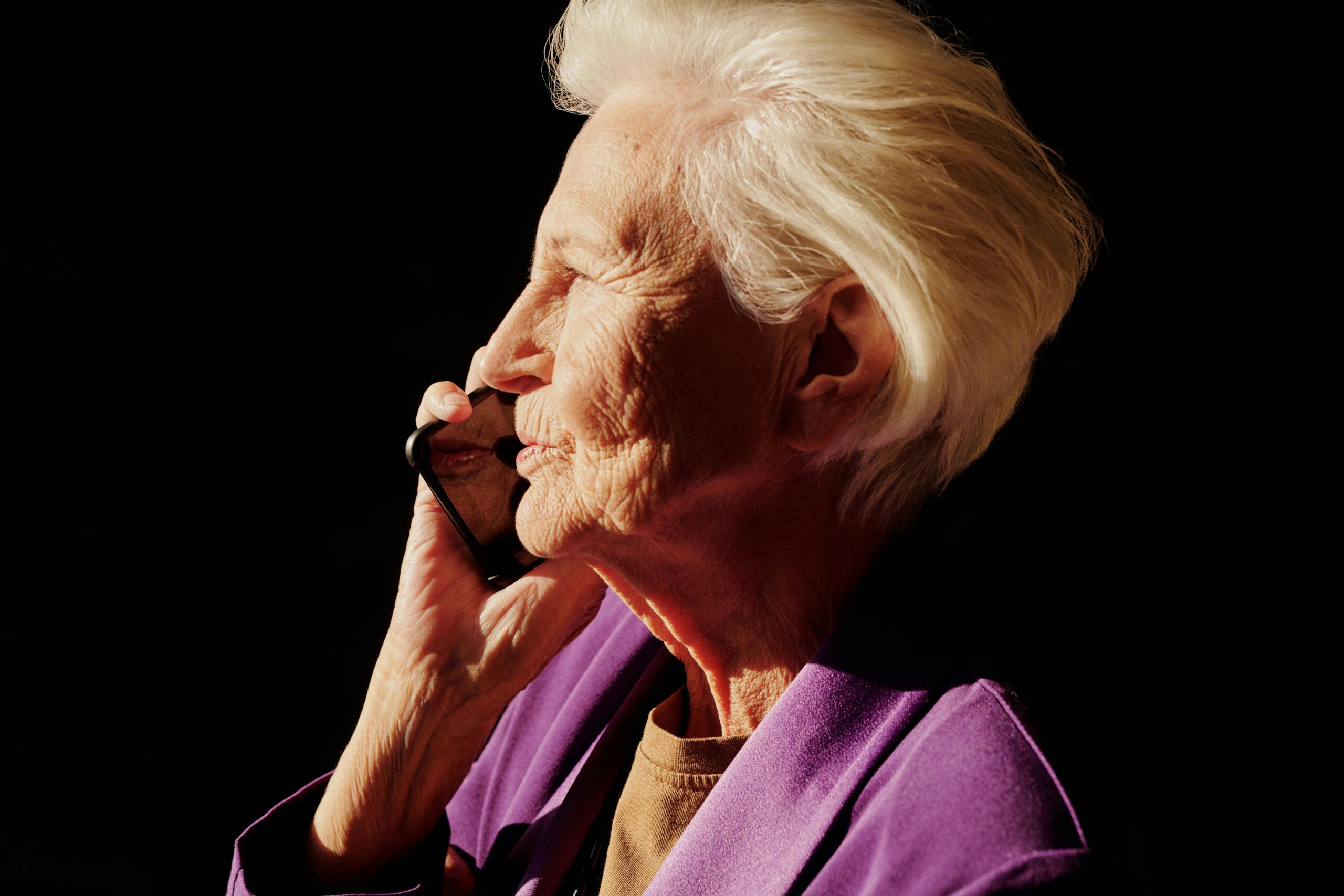 Elderly woman with white hair holding a smartphone, smiling warmly in gentle sunlight.