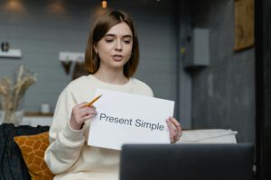 Young woman teaches English grammar online, holding a 'Present Simple' note and pencil.