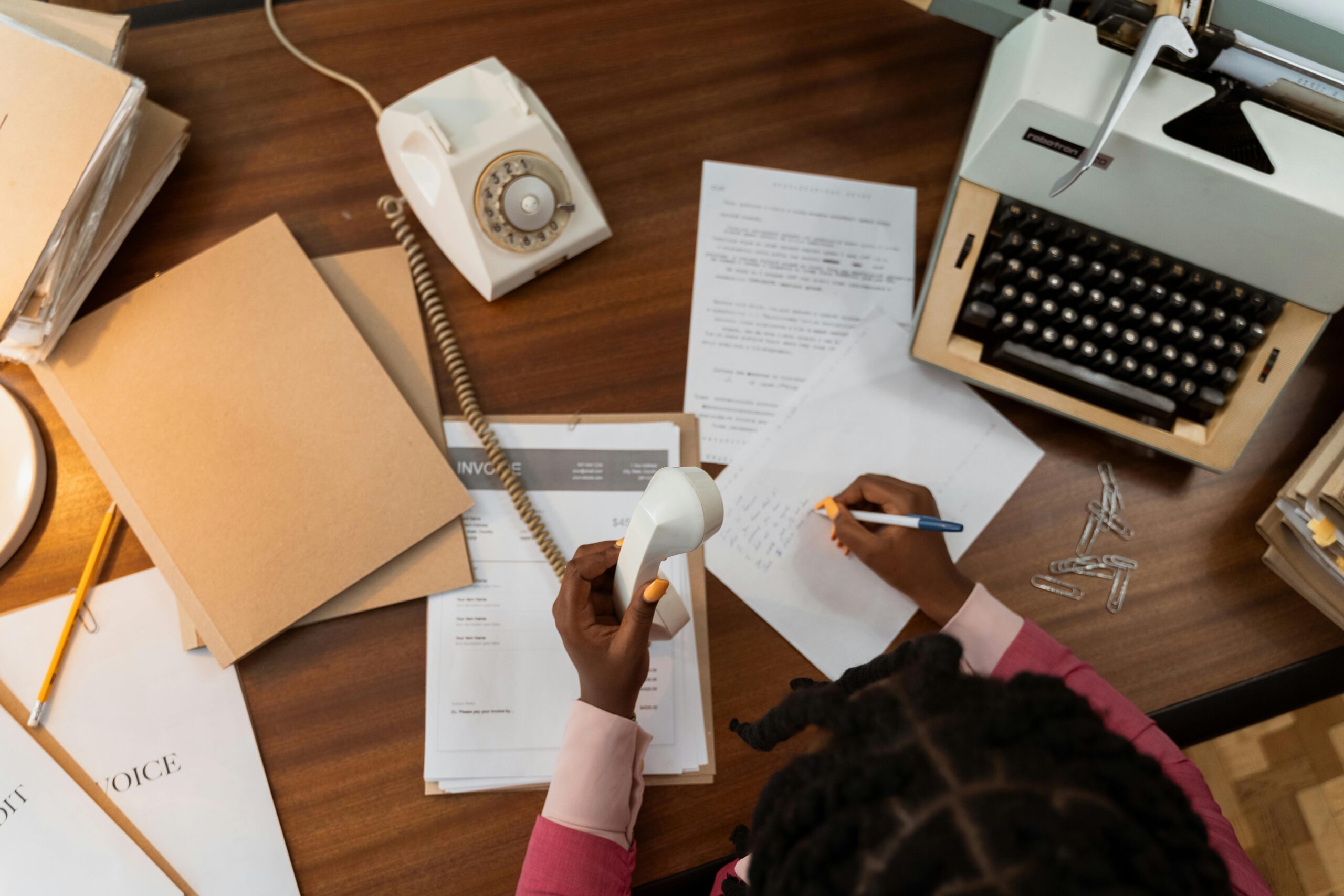 A vintage office desk with typewriter, telephone, and person writing on paper.